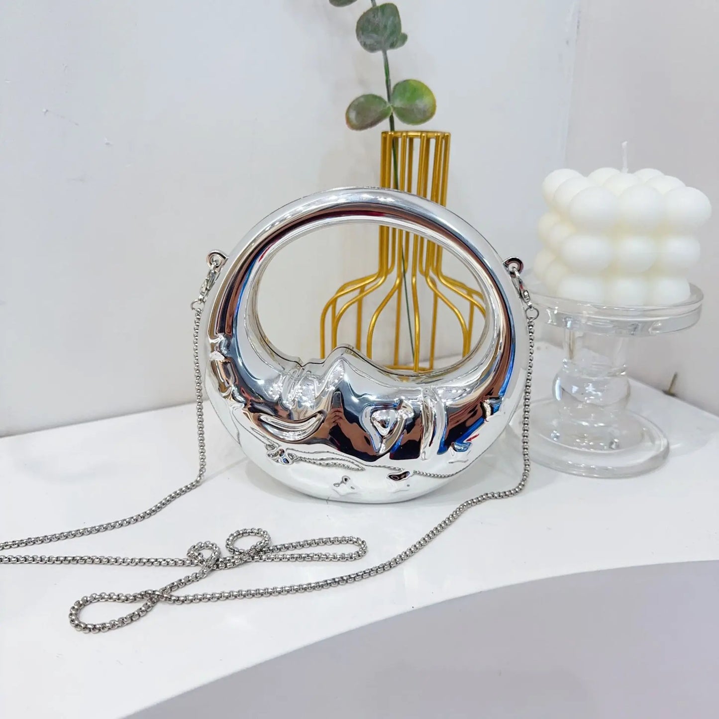 Silver crescent handbag with chain strap on a white surface with decorative items.