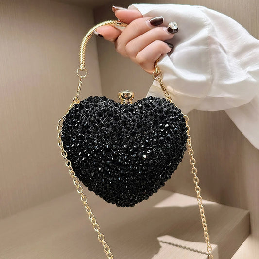 Black heart-shaped clutch bag with gold chain handle held by a hand against a neutral background