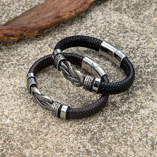 Two black braided leather bracelets with silver-toned engraved metal accents, displayed on a textured stone surface with a piece of driftwood in the background.