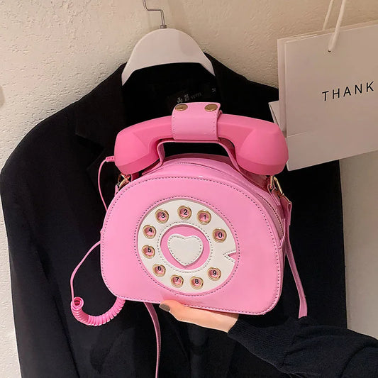 Pink phone-shaped handbag held by a person against a black garment and white wall background.