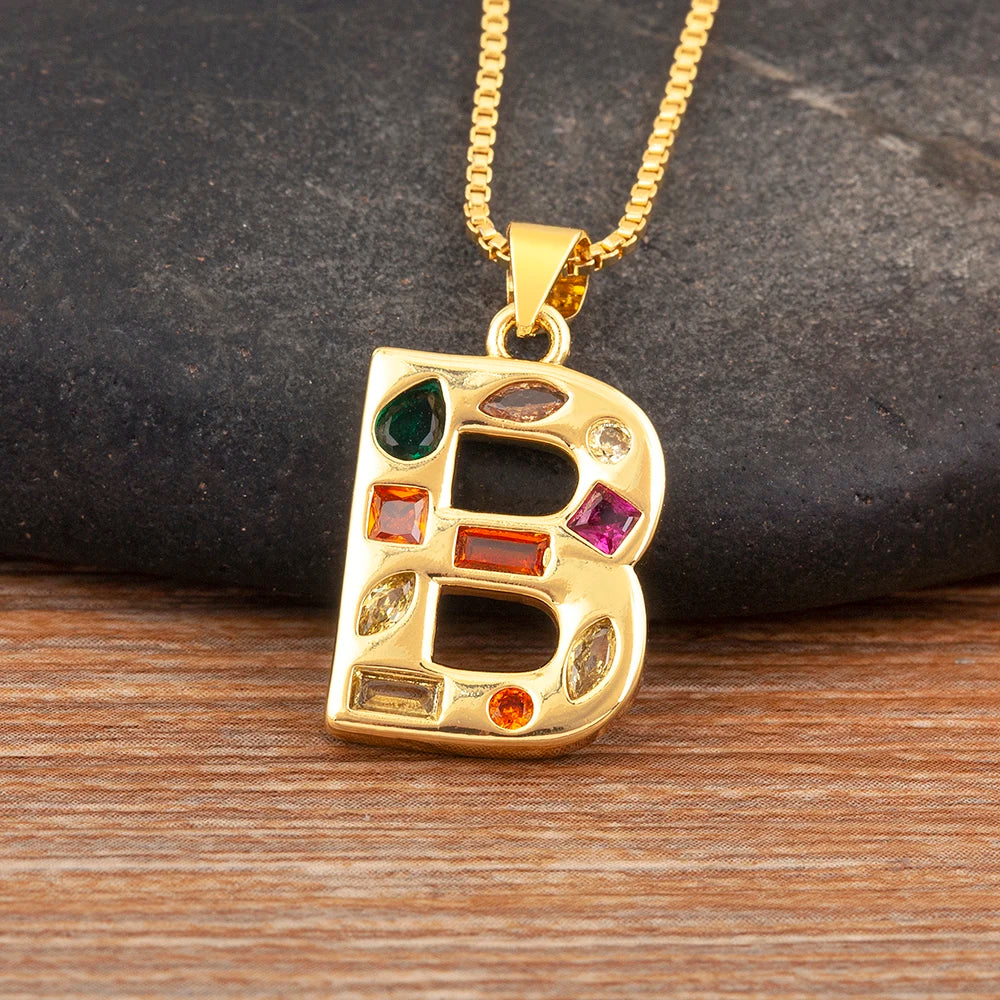 Gold necklace with a letter 'B' pendant featuring colourful gemstones on a wooden surface.