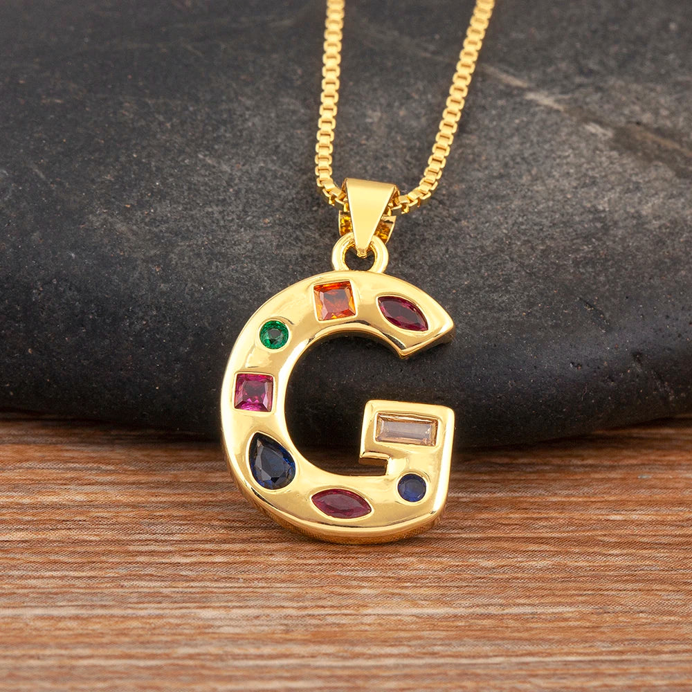 Gold necklace with a letter 'G' pendant featuring colourful gemstones on a dark stone and wooden surface.