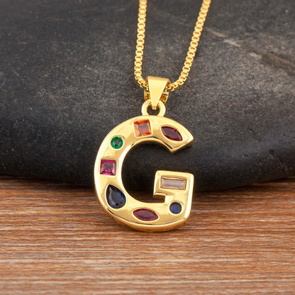 Gold necklace with a letter 'G' pendant featuring colourful gemstones on a dark stone and wooden surface.