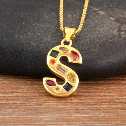Gold necklace with a letter 'S' pendant featuring colourful gemstones on a dark stone and wooden surface.