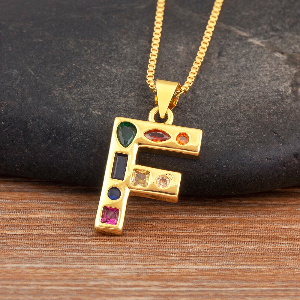 Gold necklace with a letter 'F' pendant featuring colourful gemstones on a dark stone background.