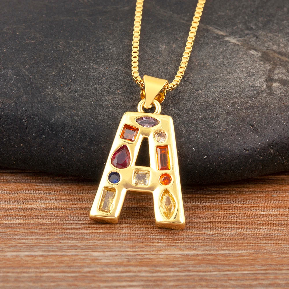 Gold necklace with a letter 'A' pendant featuring colourful gemstones on a dark stone and wooden surface.
