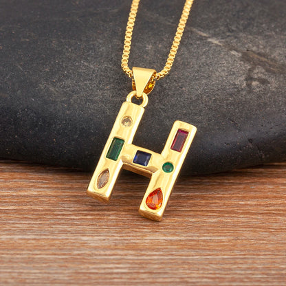 Gold necklace with a pendant shaped like the letter 'H' featuring coloured stones on a wooden surface.
