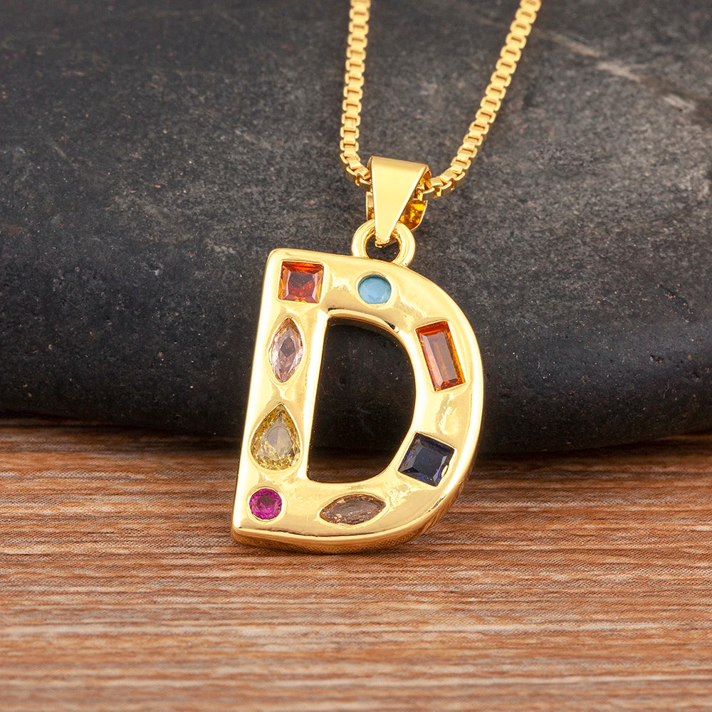 Gold necklace with a letter 'D' pendant featuring colourful gemstones on a wooden surface.