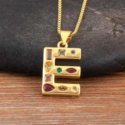 Gold necklace with a pendant shaped like the letter 'E' featuring embedded gemstones on a dark stone and wooden surface.