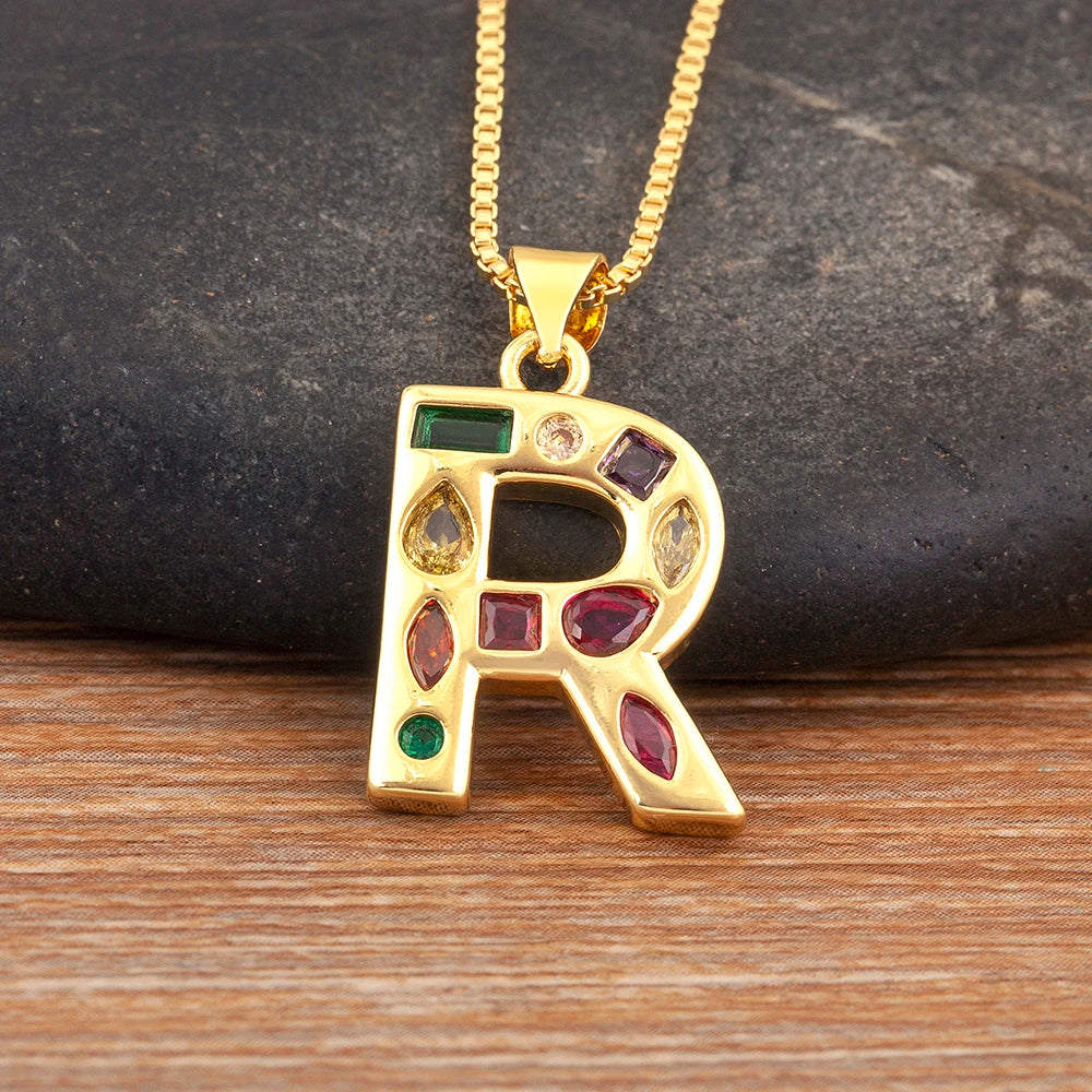 Gold necklace with a letter 'R' pendant featuring colourful gemstones on a wooden surface.