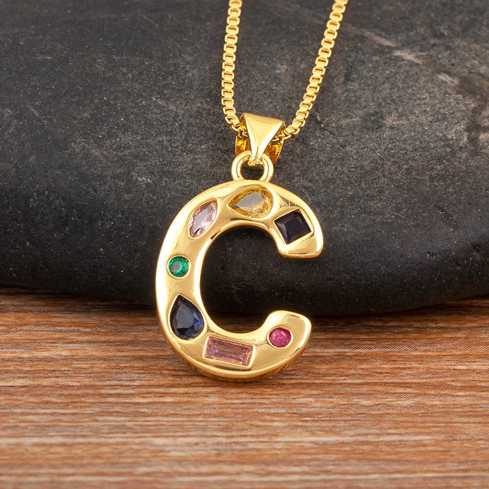 Gold necklace with a letter 'C' pendant featuring colourful gemstones on a dark stone and wooden surface.