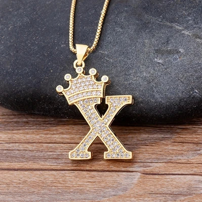 Gold necklace with a 'X' pendant featuring a crown on a dark stone and wooden surface.
