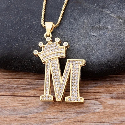 Gold necklace with a letter 'M' pendant featuring a crown on a dark stone background