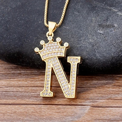Gold necklace with a letter 'N' pendant featuring a crown on a dark stone and wooden surface.