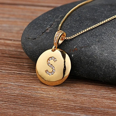 Gold necklace with a round pendant featuring the letter 'S' on a wooden surface.