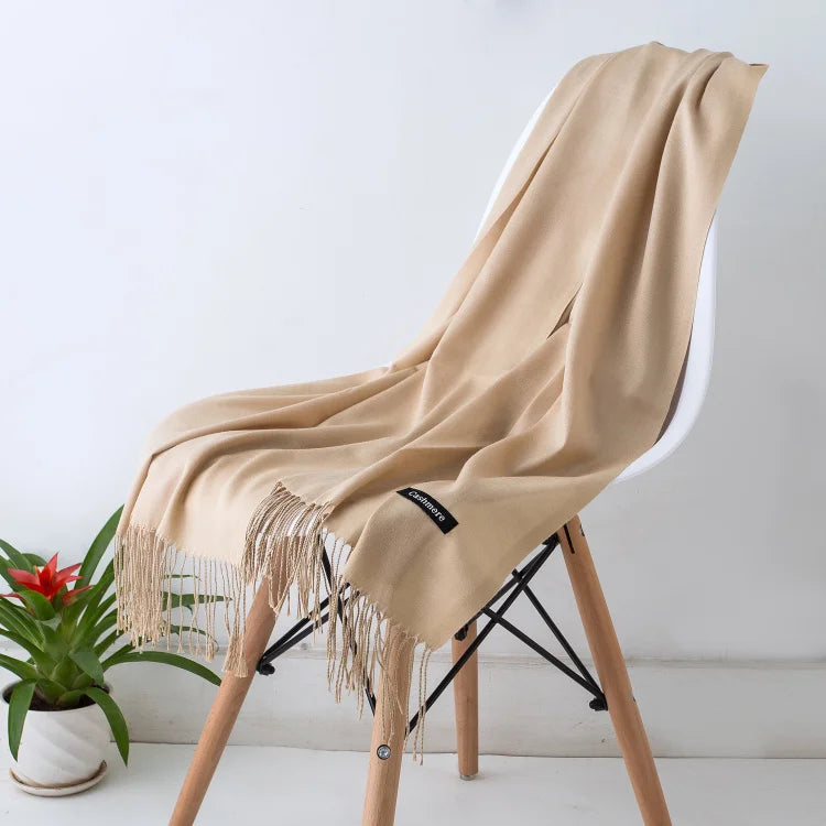 Beige scarf with fringe draped over a chair next to a plant on a white surface.