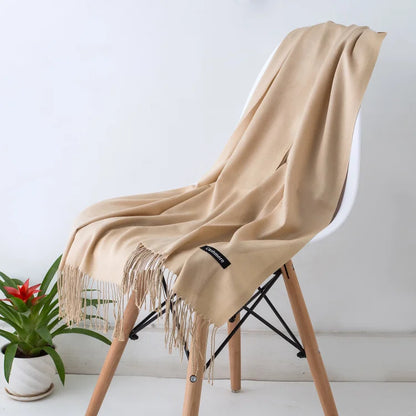 Beige scarf with fringe draped over a chair next to a plant on a white surface.