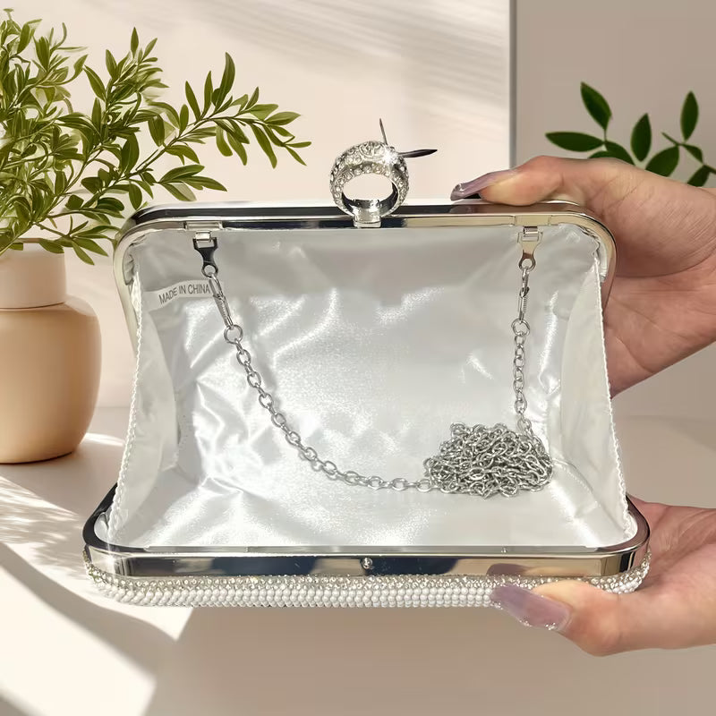 Hand holding a white clutch with silver metal chain  inside, against a neutral background with plants.