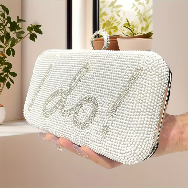 White beaded clutch with 'I do' text held by a hand, indoor setting with plants.