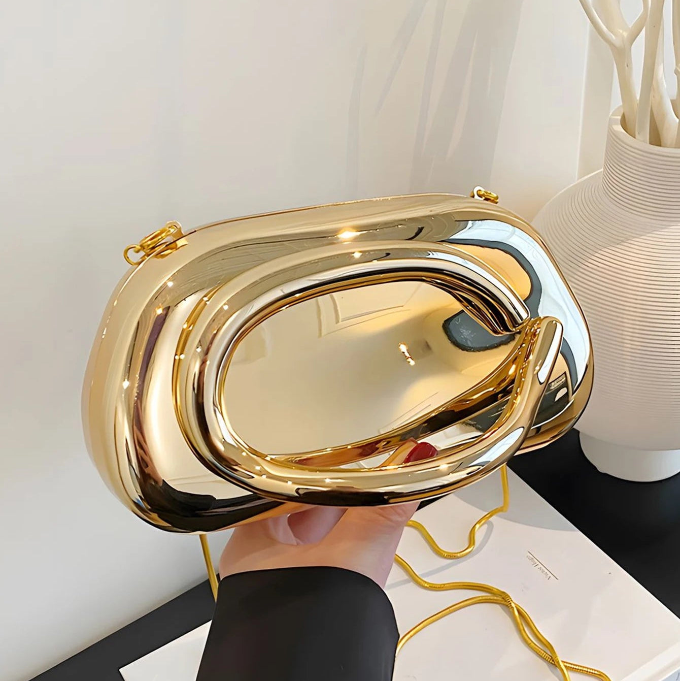 Gold clutch bag held by a hand on a white surface with a vase in the background