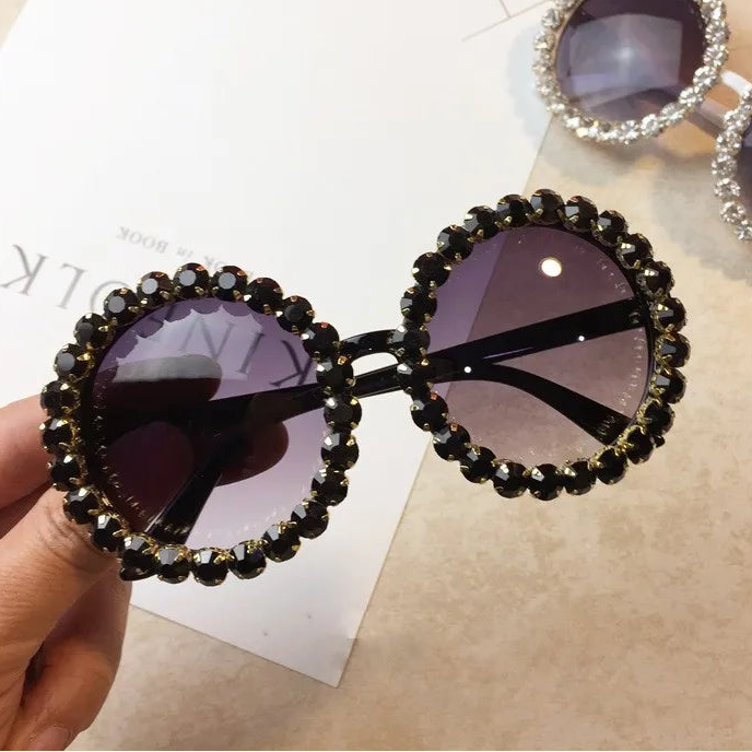Decorative sunglasses with black embellishments held by a hand on a beige surface.