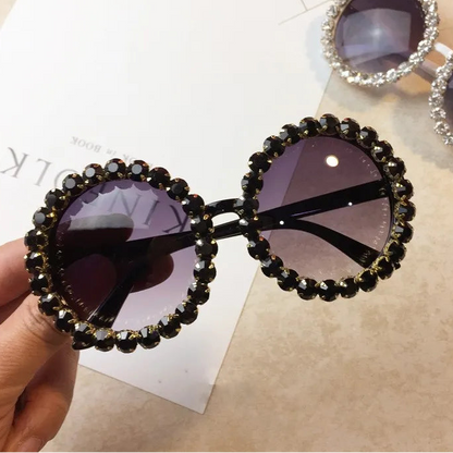 Decorative sunglasses with black embellishments held by a hand on a beige surface.