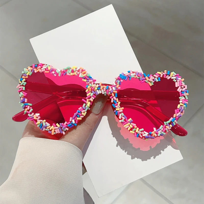 Heart-shaped sunglasses with colourful embellishments held by a hand on a light background