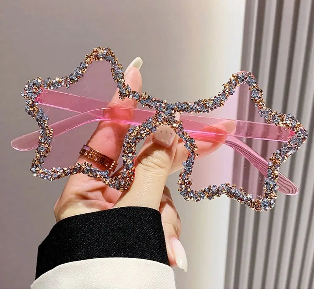 Pink star-shaped sunglasses with rhinestone frame held by a hand against a neutral background
