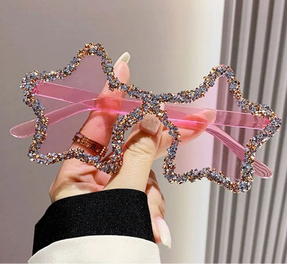 Pink star-shaped sunglasses with rhinestone frame held by a hand against a neutral background