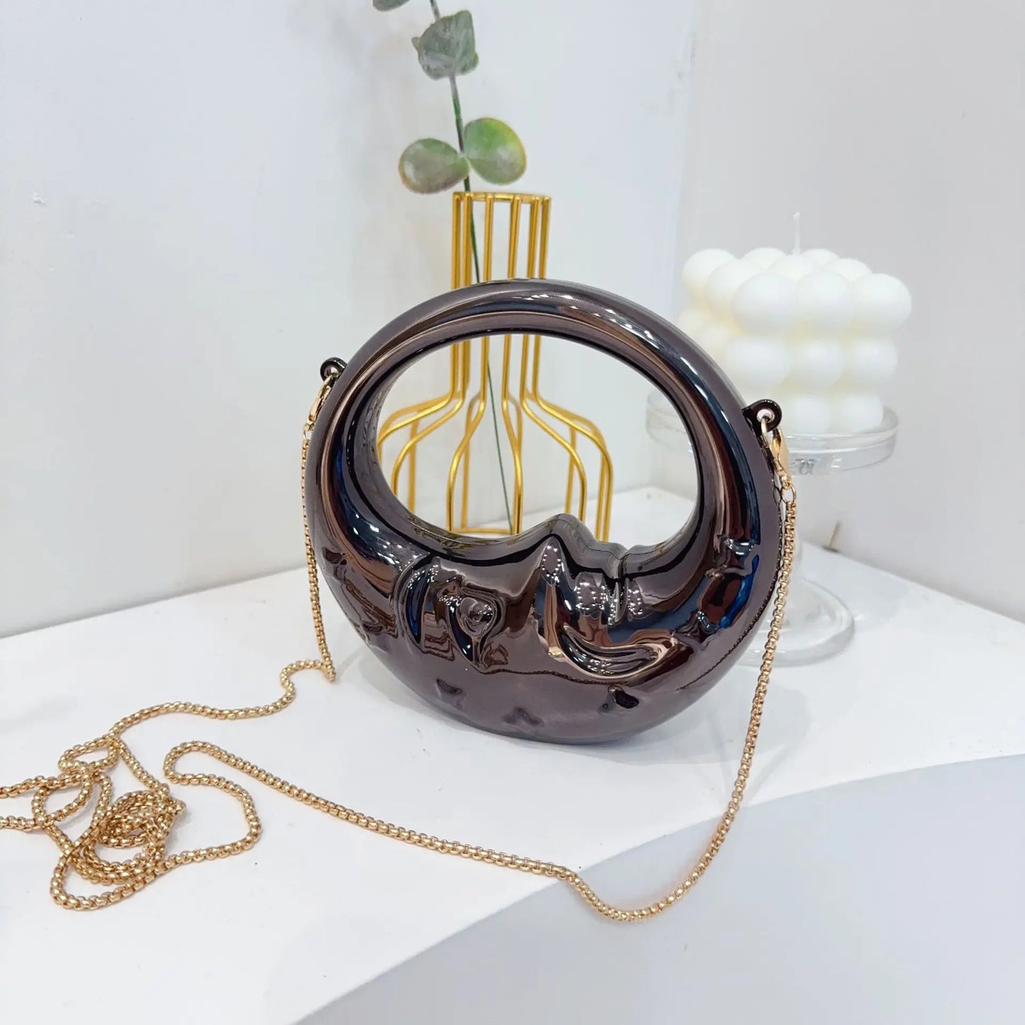 Metallic black handbag with gold chain on a white surface with decorative items in the background