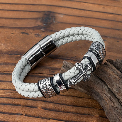 A braided light grey leather bracelet with polished silver-tone metal accents, including a cross charm and textured spacer beads, displayed on a rustic wooden surface.