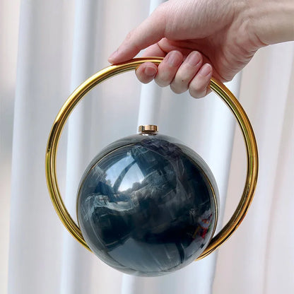 Hand holding a spherical black clutch bag with a gold handle against a white curtain background