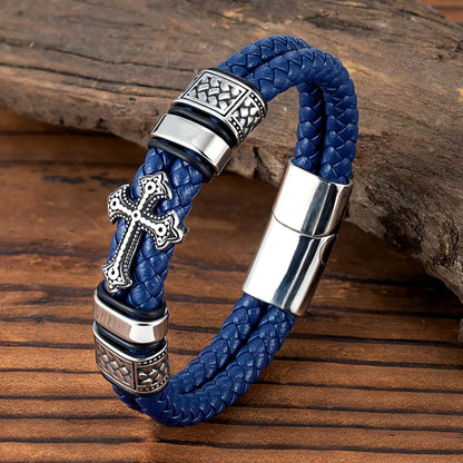 A blue braided leather bracelet with silver-tone metal accents, including a decorative cross charm, textured bezel settings, and a magnetic clasp, displayed on a rustic wooden surface.