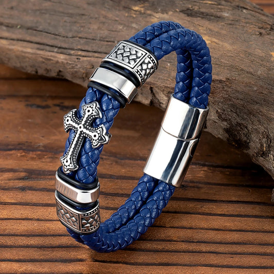 A blue braided leather bracelet with silver-tone metal accents, including a decorative cross charm, textured bezel settings, and a magnetic clasp, displayed on a rustic wooden surface.