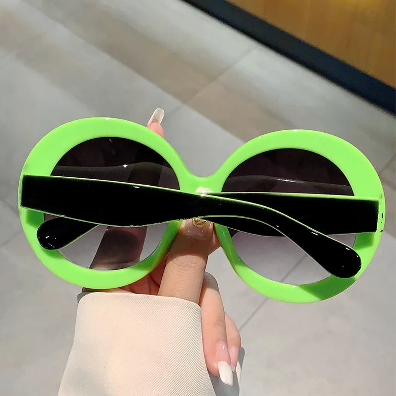 Green sunglasses with black frames held by a hand against a neutral background