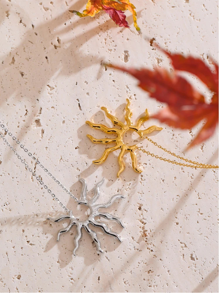Two sun-shaped pendants, one gold and one silver, on a textured surface with dried flowers.
