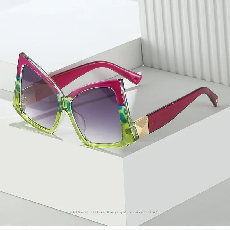 A pair of oversized square sunglasses with a translucent frame that transitions from bright lime green to vivid magenta, featuring gradient purple lenses and a gold metal hinge accent, displayed on a white geometric platform.