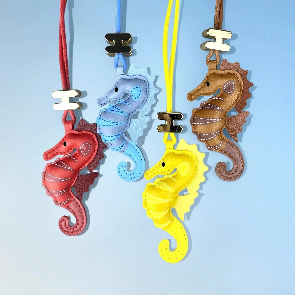 Four seahorse-shaped leather keychains in red, light blue, yellow, and brown, each hanging from a different coloured strap against a light blue background.