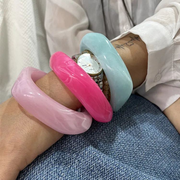 Close-up of a wrist wearing a pink and light blue bangle with a watch, on a white shirt and denim background.