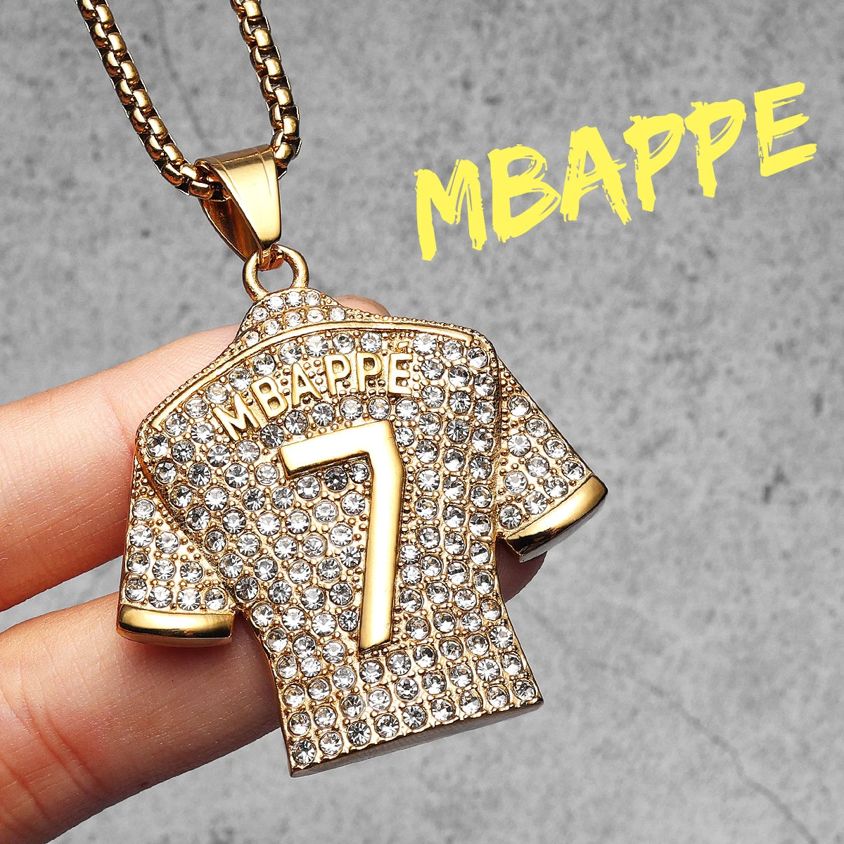 Gold pendant shaped like a sports jersey with 'MBAPPE' and number '7', held by a hand against a grey background.
