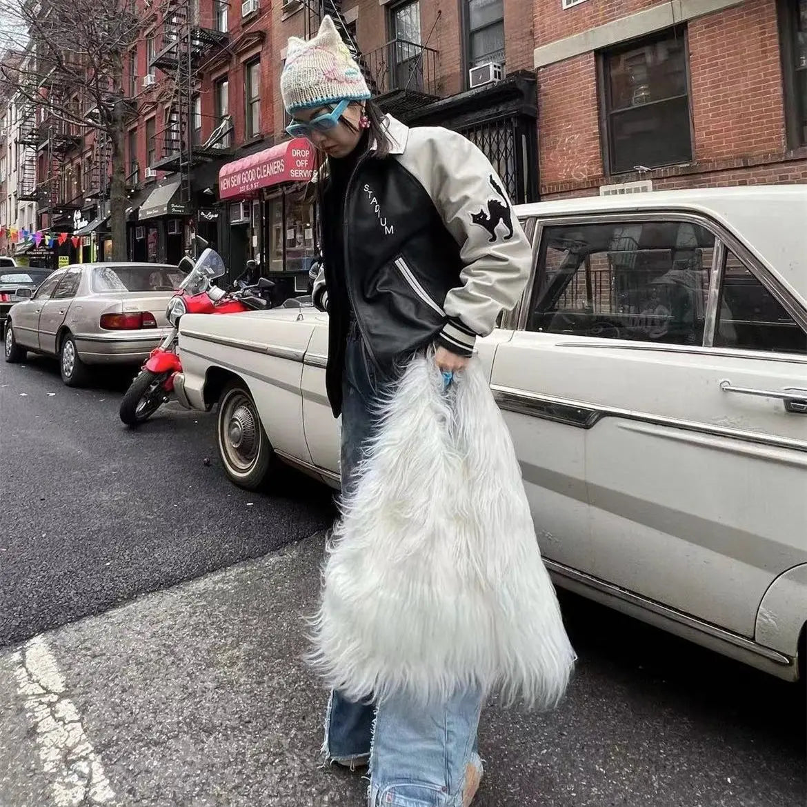 Person holding a large white fur bag on a city street with cars and buildings in the background.