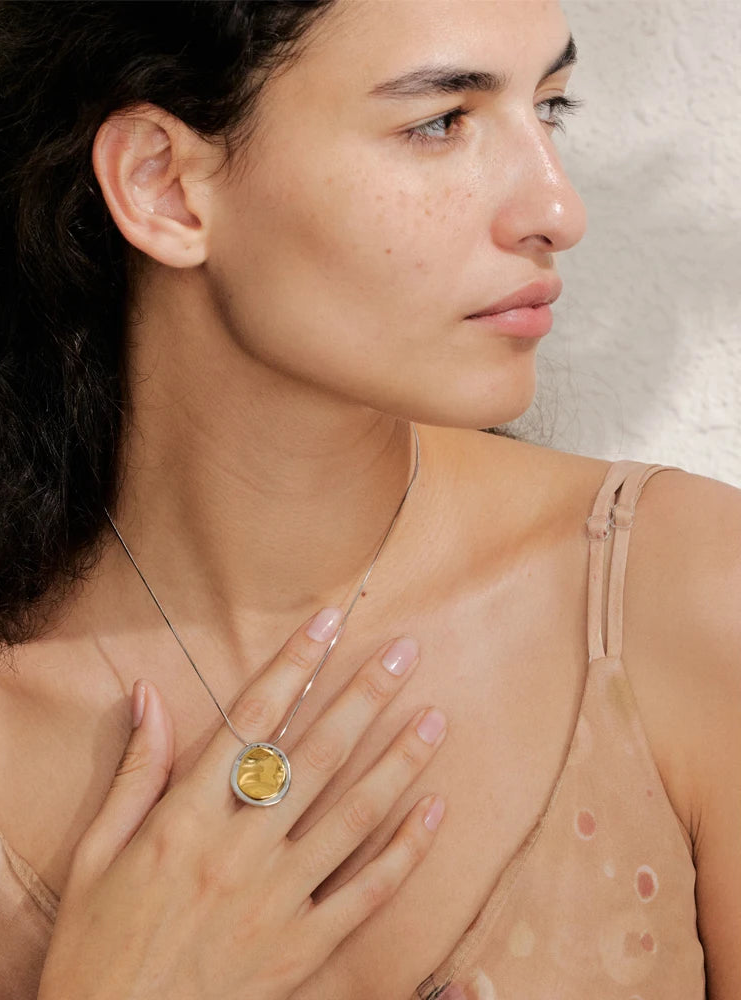 Woman wearing a gold necklace with a round pendant, holding it close to her chest.