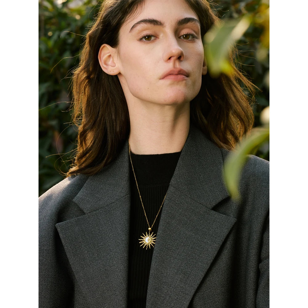 Woman wearing a grey coat and black top with a gold necklace, standing outdoors.