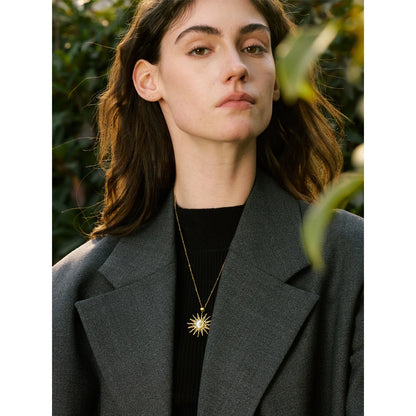 Woman wearing a grey coat and black top with a gold necklace, standing outdoors.
