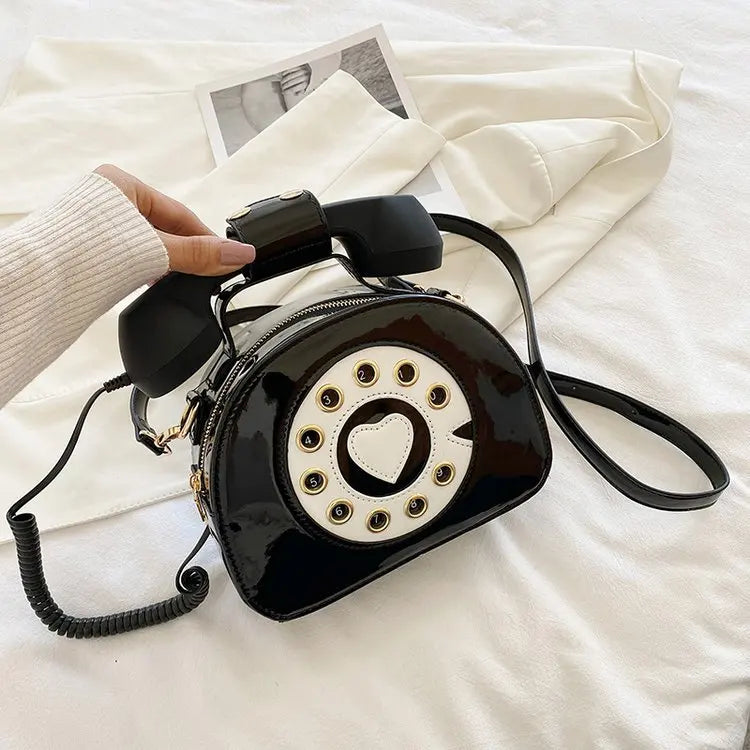 Black rotary phone-shaped handbag with a heart detail on a white surface