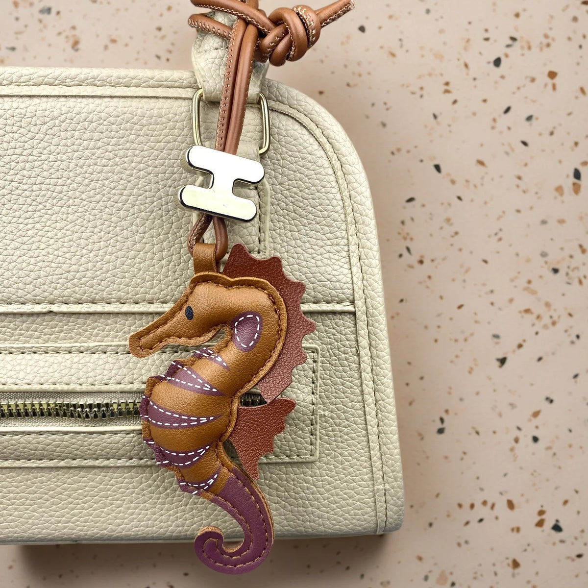 A close-up of a cream-coloured textured leather handbag featuring a brown leather seahorse keychain charm with purple accents and a silver metal 'H' charm, set against a speckled beige background.