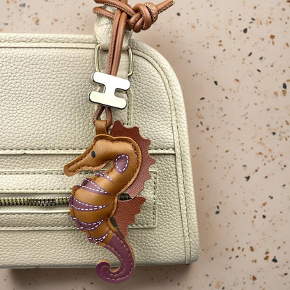 A close-up of a cream-coloured textured leather handbag featuring a brown leather seahorse keychain charm with purple accents and a silver metal 'H' charm, set against a speckled beige background.