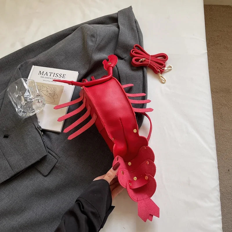 Red lobster-shaped handbag on a grey coat with a book and glass nearby.