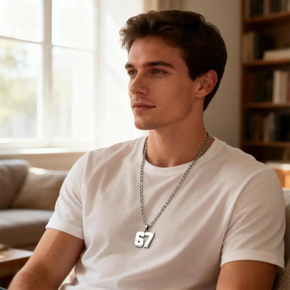 Man wearing a white t-shirt and a necklace with the number 67, sitting in a room with a window and bookshelf in the background.