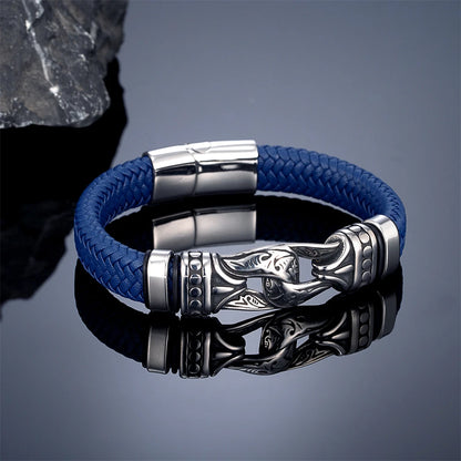 A blue braided leather bracelet with a silver-toned stainless steel magnetic clasp and a decorative engraved lobster claw connector, displayed on a reflective dark grey surface.